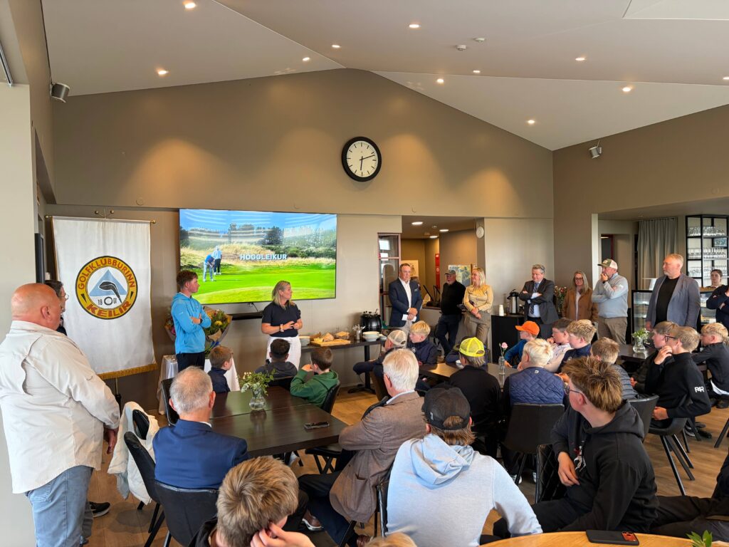 IMG_8648 - Golfsamband Íslands People gathered in a golf club meeting room listening to a presentation; a large screen shows a golf course and a wall clock is centered above the screen.