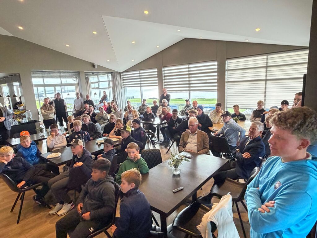 IMG_8654 - Golfsamband Íslands Crowd of children and adults seated around tables in a bright conference room with large windows overlooking water.
