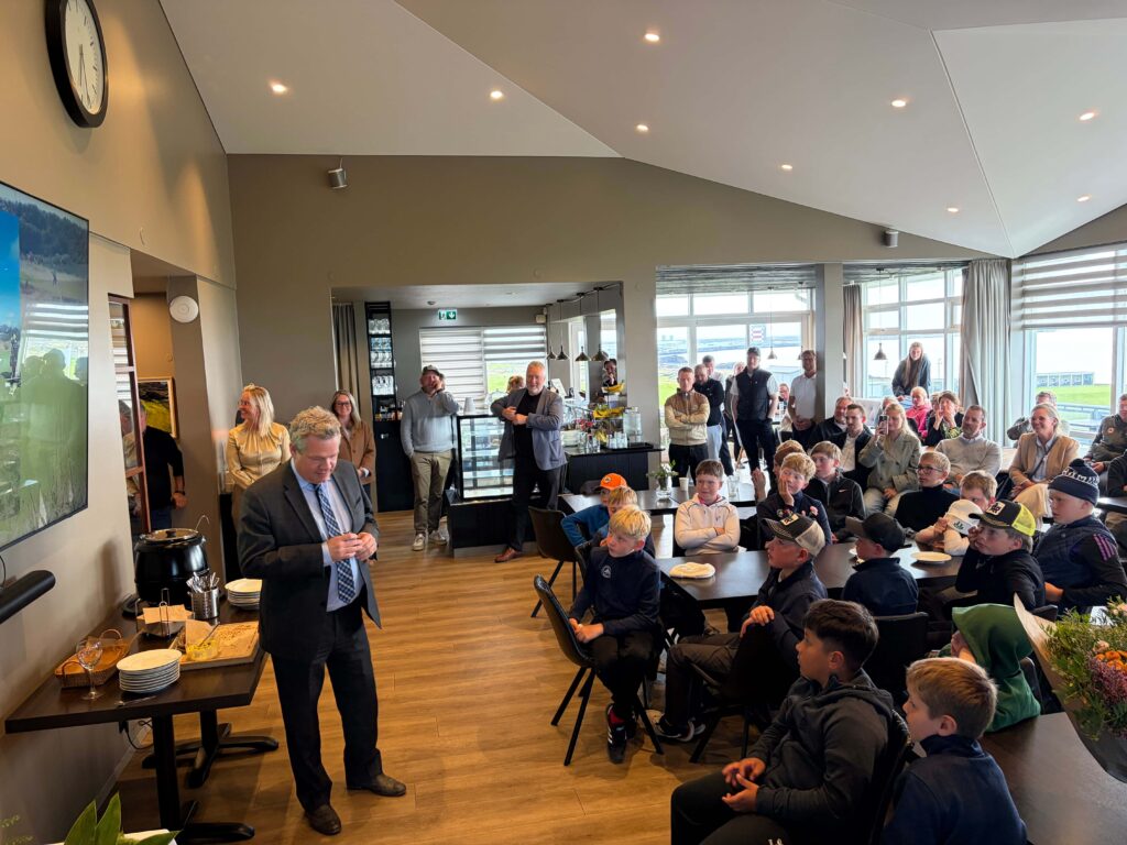 IMG_8660 - Golfsamband Íslands Man in a suit speaking to a seated audience of children and adults in a bright cafe-style room with a buffet at the side.