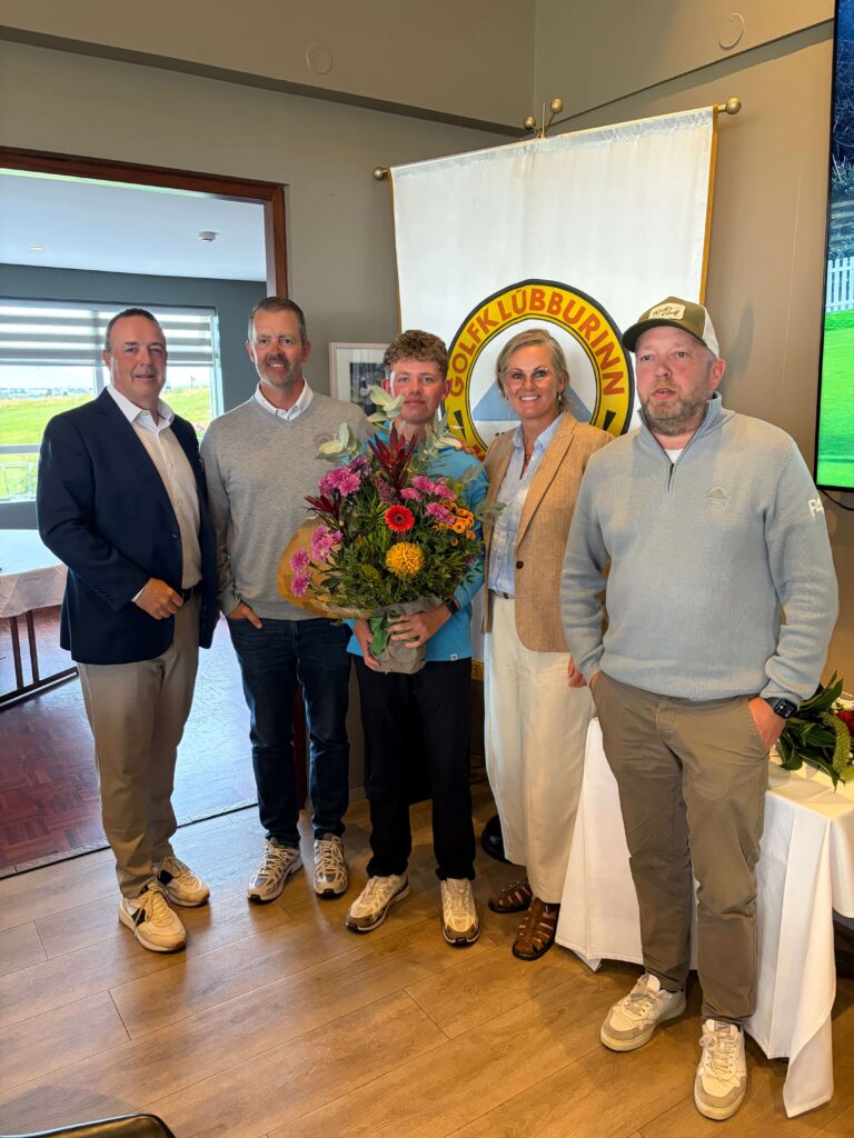 IMG_8669 - Golfsamband Íslands Five adults pose for a group photo indoors, one holding a large bouquet of colorful flowers, with a banner displaying a circular logo in the background.