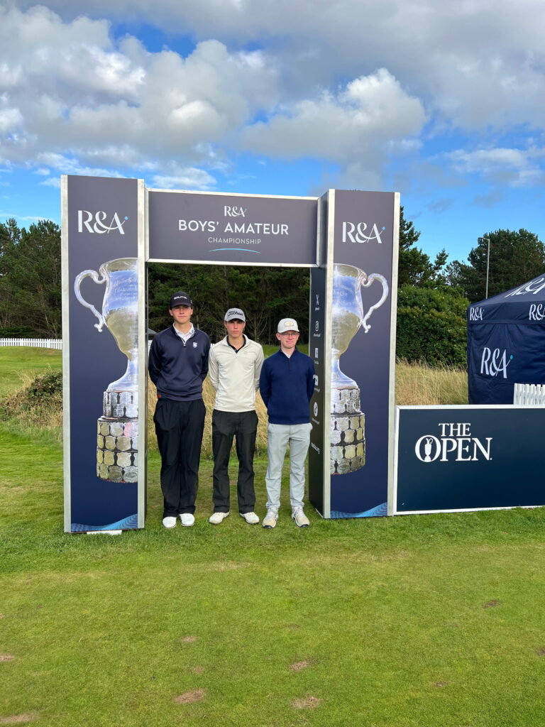 Three young golfers pose together under an R&A Boys' Amateur Championship arch with trophy graphics on the sides, blue sky behind them.