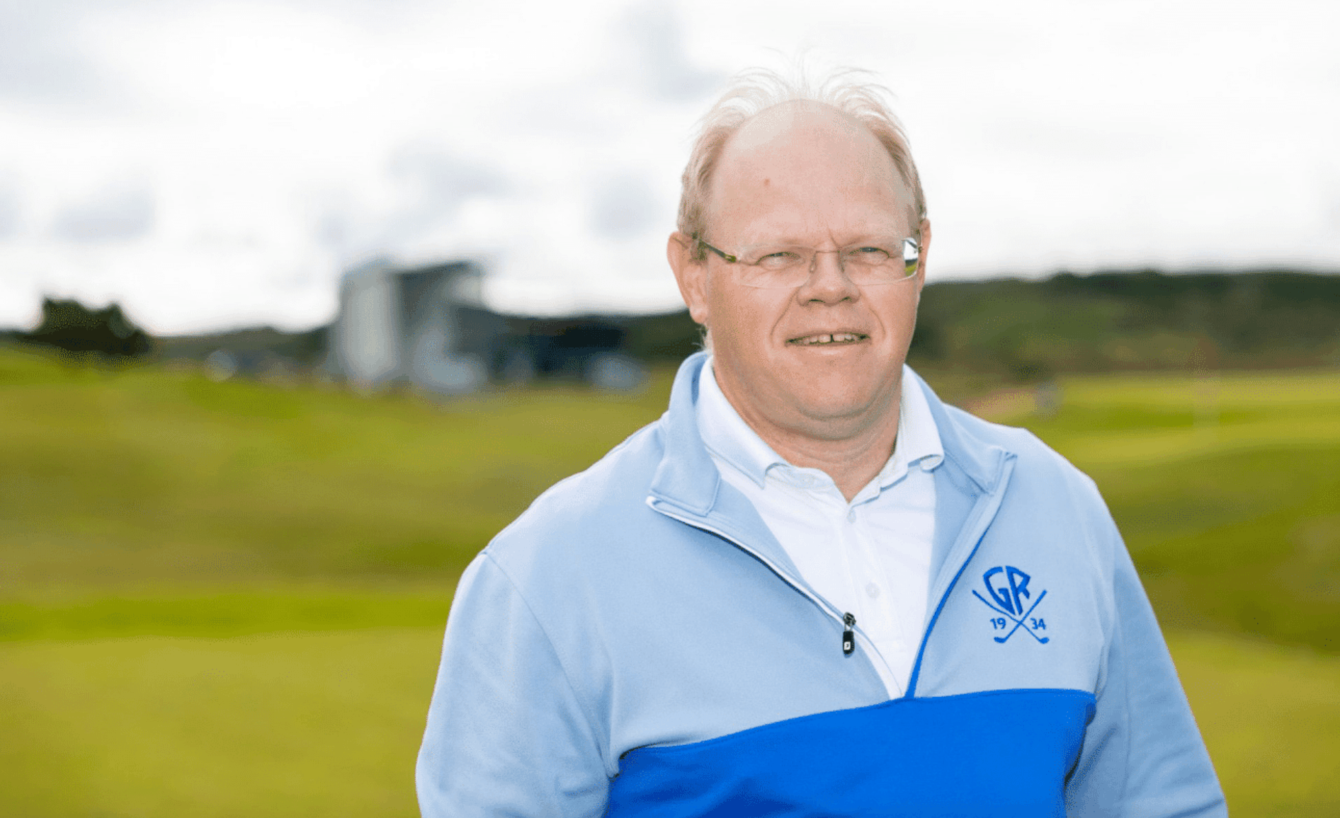 Smiling older man with glasses wearing a light blue zip-up jacket in a green field outdoors.
