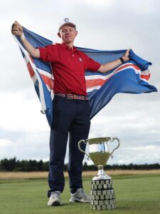 Young golfer standing on a course, arms spread with a Union Jack flag, next to a tall trophy on the grass.