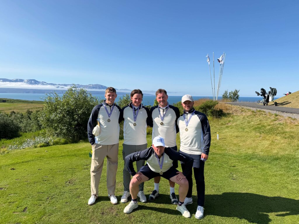f7e1c129-7f69-4797-a01f-45e3bce994f9 - Golfsamband Íslands Four men in matching athletic tops posing with medals on a sunny coastal golf course, with blue sky and distant mountains.