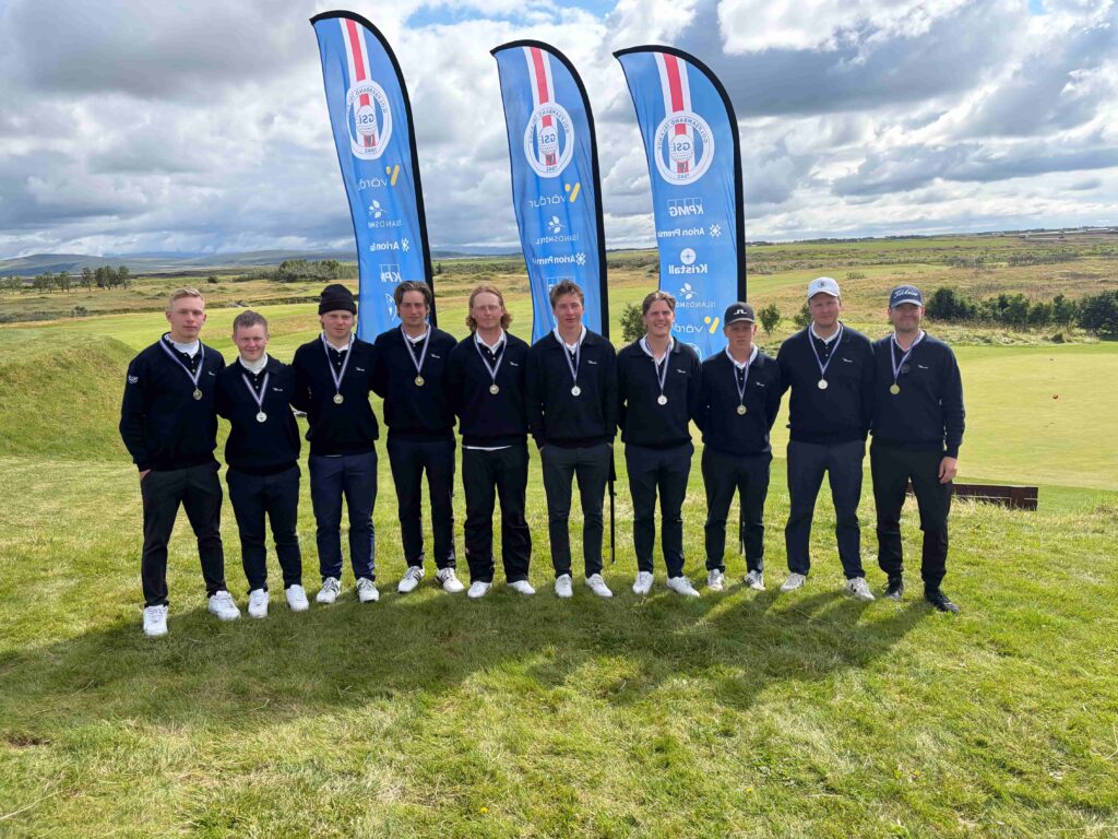A group of young men wearing dark sweaters and medals posing for a photo on a grassy golf course with tall blue sponsor flags in the background.