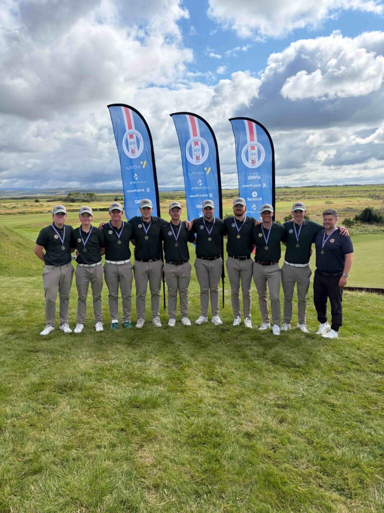 Eleven men in dark polo shirts and caps with medals pose on a golf course, arms around each other, with tall blue sponsor flags in the background and a cloudy sky above.