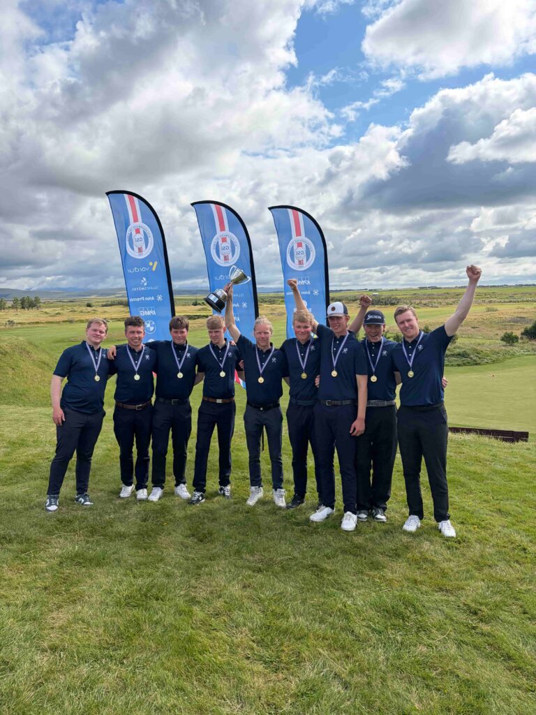 Group of young men in navy polo shirts with medals, posing on a golf course with a trophy and blue sponsor flags behind them.