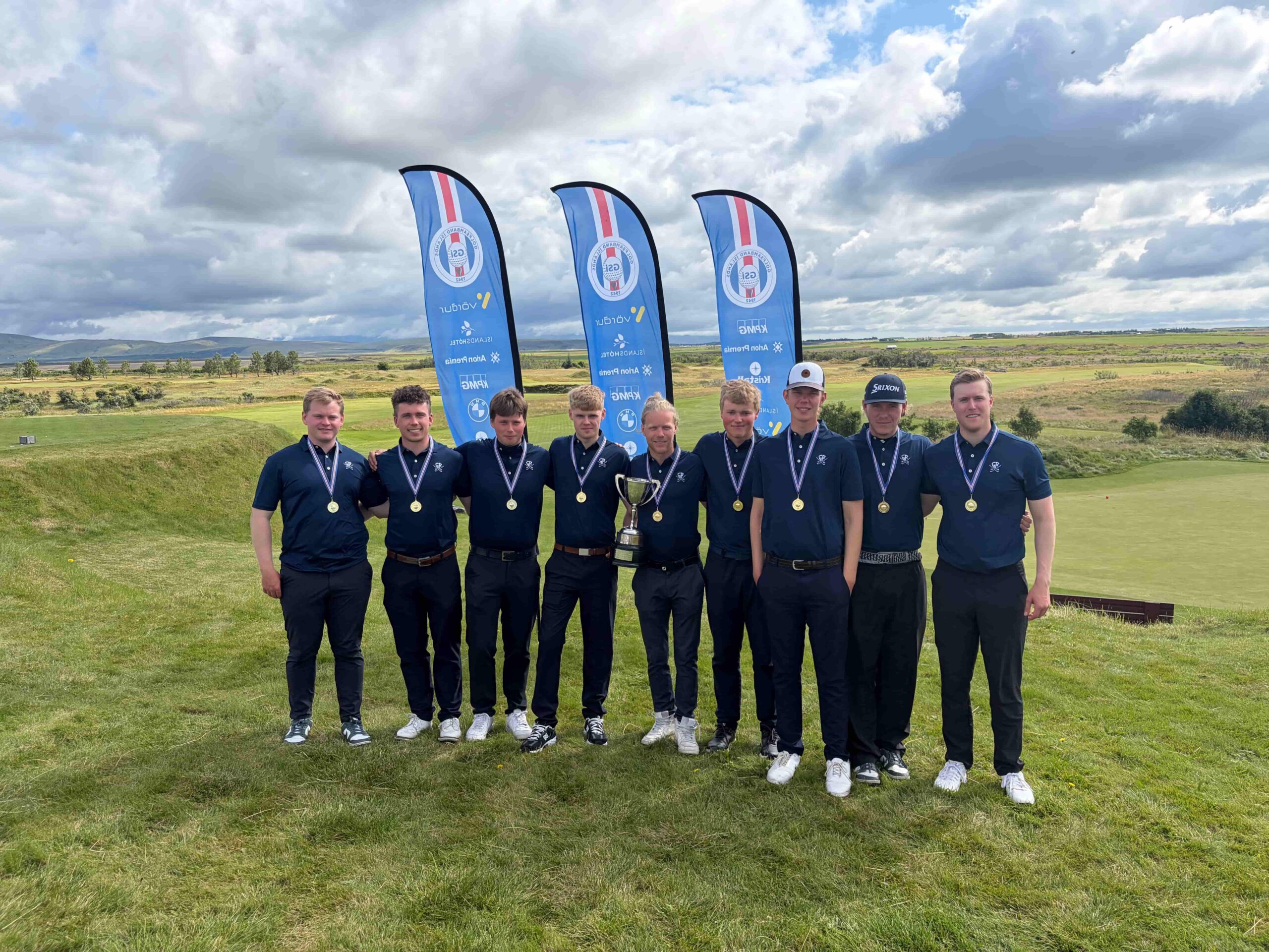 A group of young men in navy polo shirts with gold medals around their necks, posing outdoors with a trophy on a golf course, blue flags in the background.