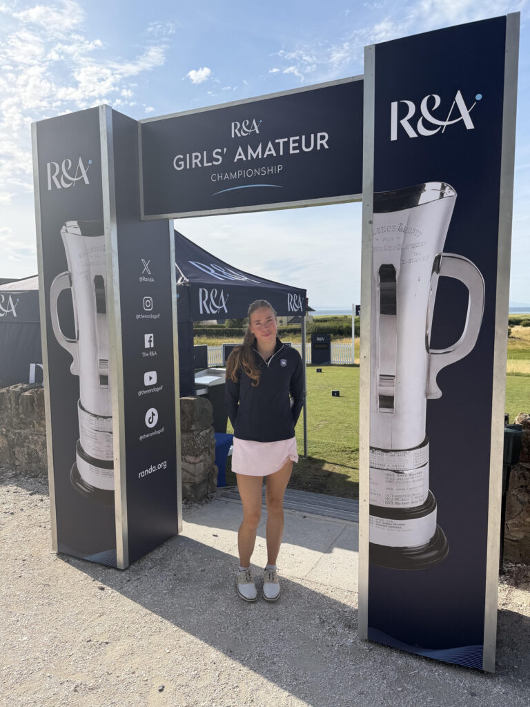 unnamed 44 - Golfsamband Íslands Young woman posing under an R&A Girls' Amateur Championship sign at an outdoor golf event.