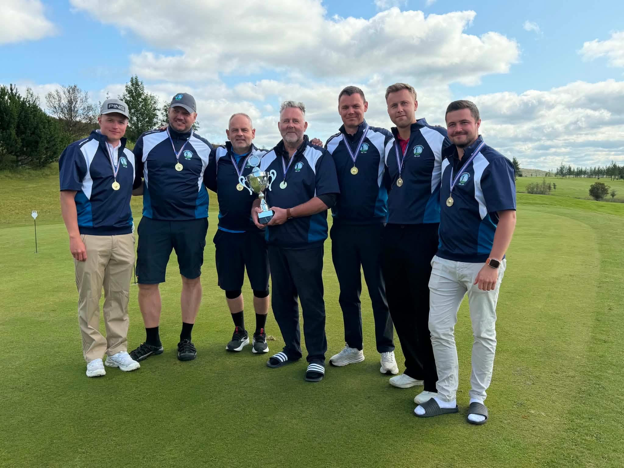 Seven golfers in matching blue uniforms with medals, posing together on a golf course with a trophy in the center.