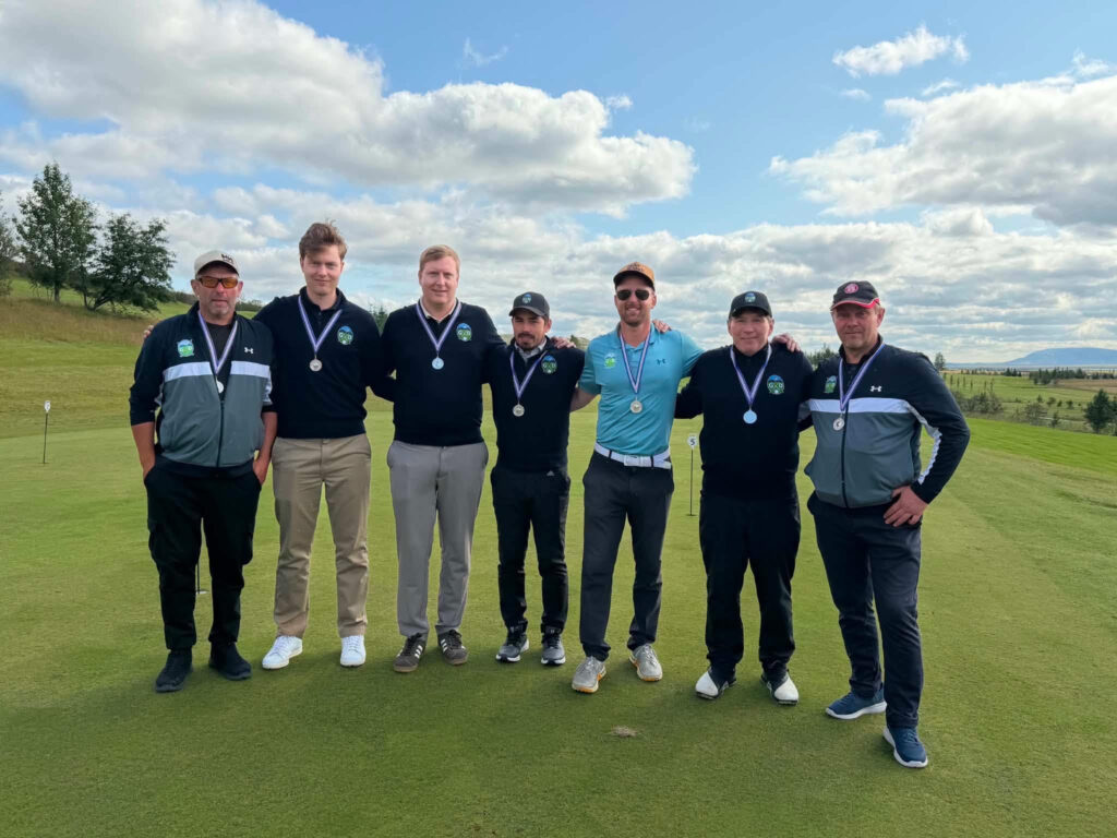 unnamed 46 - Golfsamband Íslands Seven men stand side by side on a golf course, each wearing medals, arms around shoulders, under a blue sky.