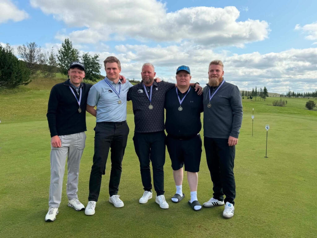 unnamed 47 - Golfsamband Íslands Five men stand together on a golf course, each wearing medals around their necks and smiling with arms around each other in a sunny, grassy landscape.